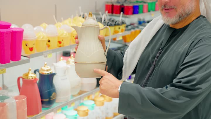 التسوق داخل المتاجر التجارية ، تصوير لرجل عربي خليجي مسن يرتدي الكندورة و الغترة يتفقد دلال القهوة العربية داخل متجر أدوات منزلية  و يشير اليها، الاهتمام بتجهيز مستلزمات الضيافة العربية