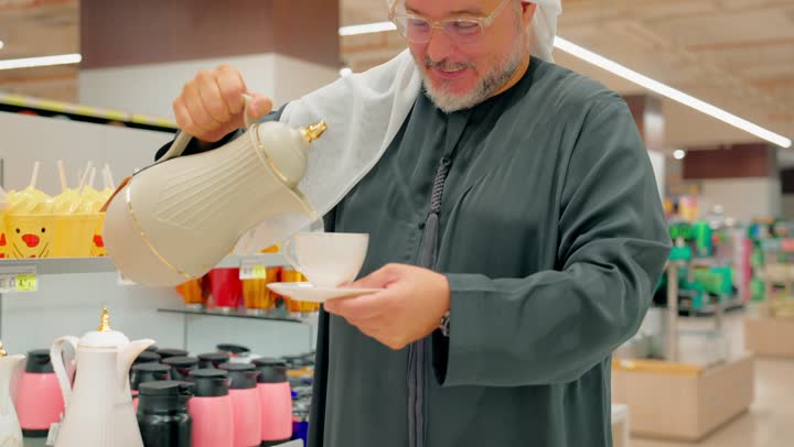 الاهتمام بالتقاليد العربية في الضيافة، رجل عربي خليجي اماراتي مسن يرتدي الكندورة و الغتره يتفقد أدوات الضيافة داخل متجر أدوات منزلية يقوم بسكب القهوة باستخدام دلة وفنجان عربي، اختيار أدوات التقديم المناسبة للضيافة العربية