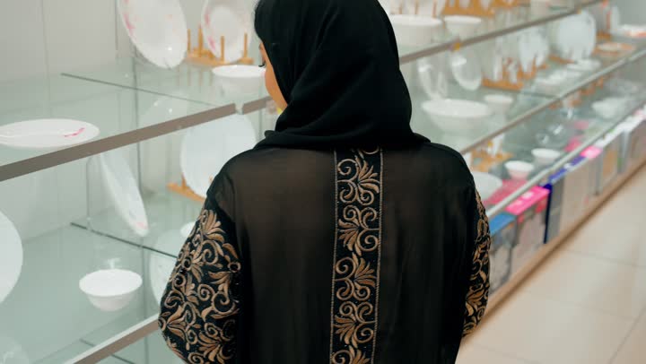 Choosing kitchen supplies and household tools, a close-up shot from behind of a veiled Emirati Gulf Arab woman inspecting household items inside the store.