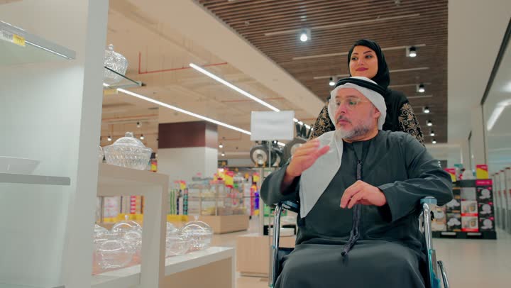 اختيار ومقارنة الاطباق أثناء التسوق، رجل مسن عربي خليجي اماراتي يرتدي الزي التقليدي يجلس على كرسي متحرك برفقة ابنته، الاهتمام برعاية كبار السن داخل المتجر، قضاء وقت ممتع في شراء الحاجيات المنزلية