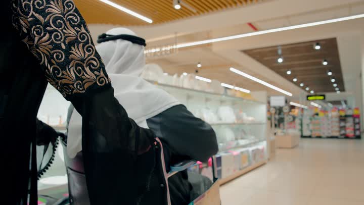 الاشارة الى شيء ما، تصوير مقرب لرجل مسن عربي خليجي اماراتي يرتدي الزي التقليدي يجلس على كرسي متحرك داخل متجر أدوات منزلية برفقة ابنته، الاهتمام برعاية كبار السن داخل المتجر، قضاء وقت ممتع في شراء الحاجيات المنزلية