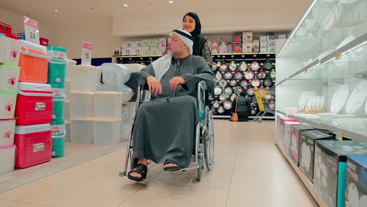 رجل مسن عربي خليجي اماراتي يرتدي الزي التقليدي يجلس على كرسي متحرك داخل متجر أدوات منزلية برفقة ابنته، الاهتمام برعاية كبار السن داخل المتجر، قضاء وقت ممتع في شراء الحاجيات المنزلية