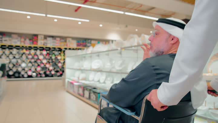 الاهتمام براحة كبار السن أثناء التسوق، التعاون الأسري داخل السوبرماركت، رجل عربي خليجي اماراتي مسن يرتدي الزي التقليدي يجلس على كرسي متحرك داخل متجر أدوات منزلية