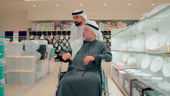 التعاون الأسري داخل السوبرماركت، رجل عربي خليجي اماراتي مسن يرتدي الزي التقليدي يجلس على كرسي متحرك داخل متجر أدوات منزلية ، الاهتمام براحة كبار السن أثناء التسوق