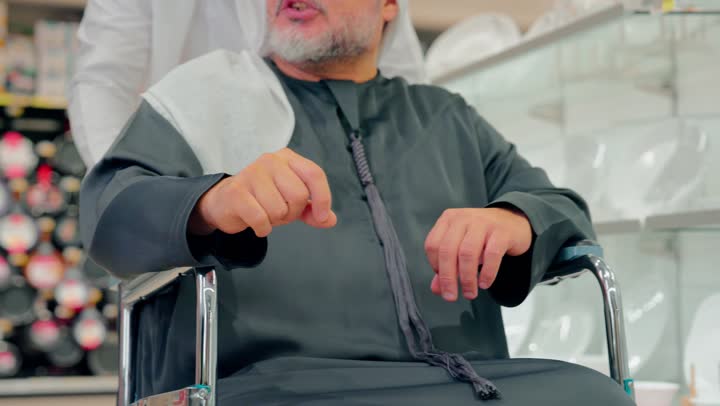 تصوير مقرب لرجل عربي خليجي اماراتي يرتدي الزي التقليدي يدفع رجل مسن يجلس على كرسي متحرك داخل متجر حديث، دعم كبار السن أثناء التسوق، الاهتمام براحة المسنين أثناء التنقل