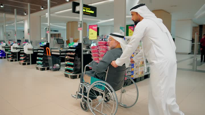 Gestures of happiness and joy, supporting the elderly while shopping, caring for the comfort of seniors while moving, an Emirati Gulf Arab man wearing traditional attire pushing an elderly man sitting in a wheelchair inside a modern store.