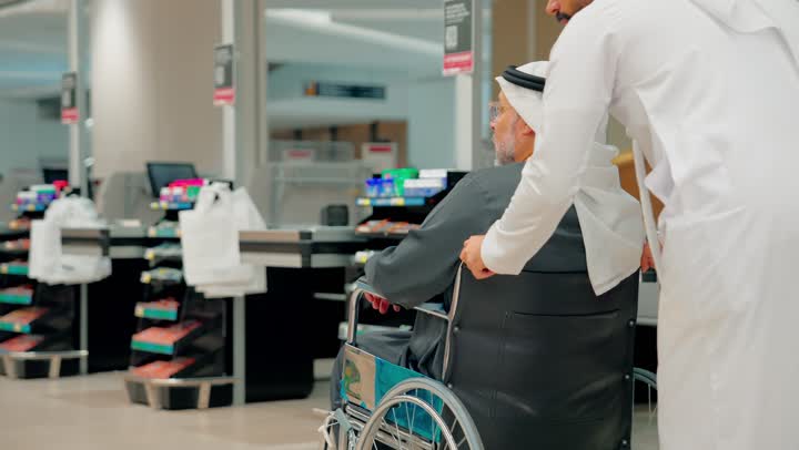 دعم كبار السن أثناء التسوق، الاهتمام براحة المسنين أثناء التنقل، رجل عربي خليجي اماراتي يرتدي الزي التقليدي يدفع رجل مسن يجلس على كرسي متحرك داخل متجر حديث، ايماءات السعادة و السرور