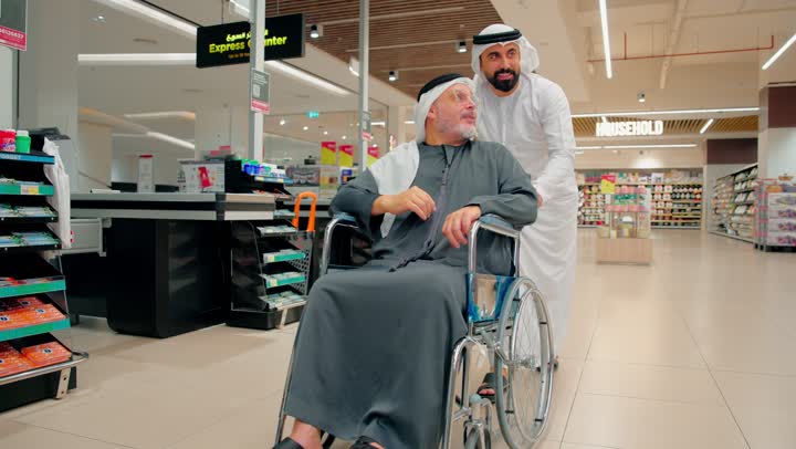 Caring for the comfort of the elderly while moving, an Arab Gulf Emirati man wearing traditional attire is pushing an elderly man sitting in a wheelchair inside a modern store, supporting the elderly while shopping, gestures of happiness and joy.