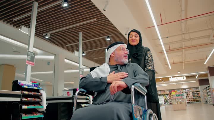التسوق داخل المراكز التجارية الحديثة، رجل عربي خليجي اماراتي مسن يرتدي الزي التقليدي يجلس على كرسي متحرك داخل متجر حديث برفقة ابنته، الاهتمام بكبار السن داخل الأماكن العامة، دعم الأسرة والرعاية الإنسانية