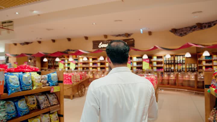 Individual shopping inside retail stores, carefully selecting daily food products, an Arab Gulf Emirati man dressed in casual attire wandering inside a modern supermarket, checking the shelves of snacks and various sweets.