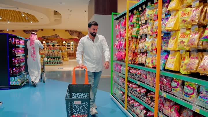 اختيار المنتجات الغذائية اليومية، رجل عربي خليجي اماراتي يرتدي الزي الكاجوال يتجول داخل سوبر ماركت حديث، تفقد أرفف السناكس والحلويات المتنوعة، التسوق الفردي داخل المتاجر التجارية