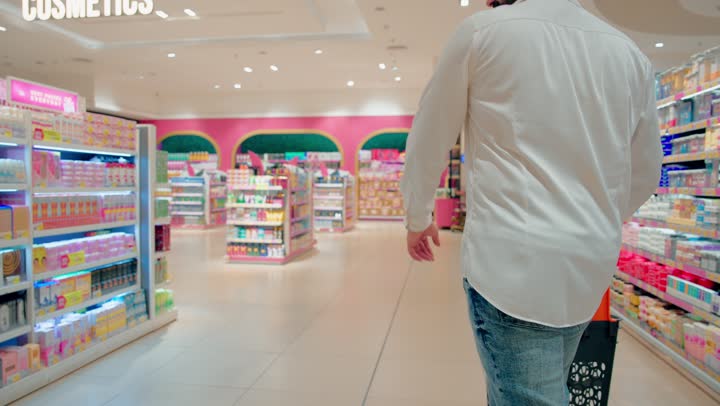 اختيار منتجات العناية الشخصية اليومية، تصوير من الخلف لرجل عربي خليجي اماراتي يرتدي الزي الكاجوال يمسك بيدية سله التسوق يتجول داخل متجر مستحضرات تجميل ومنتجات عناية شخصية