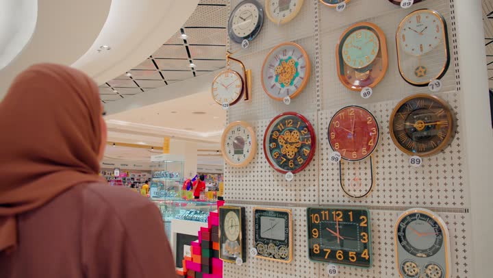 الاهتمام بالديكور المنزلي العصري، مفهوم اختيار الوقت وتنظيم المنزل، امرأة عربية خليجيه اماراتية محجبة تقوم باختيار ساعات حائط متنوعة داخل المتجر ، تشكيلة مختلفة من ساعات الحائط بتصاميم وألوان متعددة، التسوق داخل المراكز التجارية الحديثة