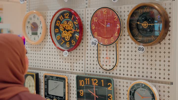 مفهوم اختيار الوقت وتنظيم المنزل، امرأة عربية خليجيه اماراتية محجبة تقوم باختيار ساعات حائط متنوعة داخل المتجر ، تشكيلة مختلفة من ساعات الحائط بتصاميم وألوان متعددة، التسوق داخل المراكز التجارية الحديثة، الاهتمام بالديكور المنزلي العصري