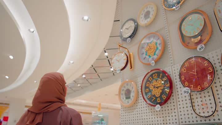 امرأة عربية خليجيه اماراتية محجبة تقوم باختيار ساعات حائط متنوعة داخل المتجر ، تشكيلة مختلفة من ساعات الحائط بتصاميم وألوان متعددة، مفهوم اختيار الوقت وتنظيم المنزل، التسوق داخل المراكز التجارية الحديثة، الاهتمام بالديكور المنزلي العصري