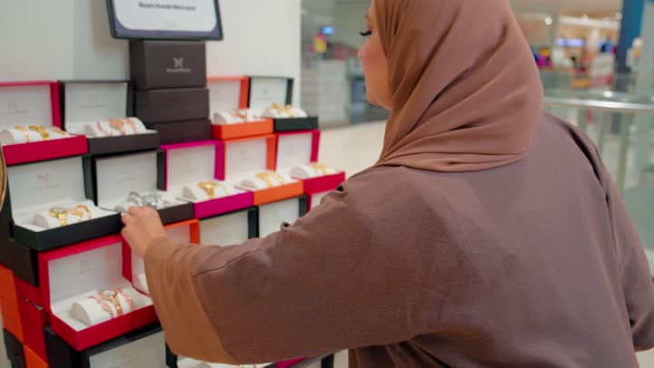 Studying options before completing a purchase, the concept of conscious shopping, an Arab Gulf Emirati woman holding a luxury watch with an elegant design, inspecting it among the watch products inside the store, attention to detail and product quality.