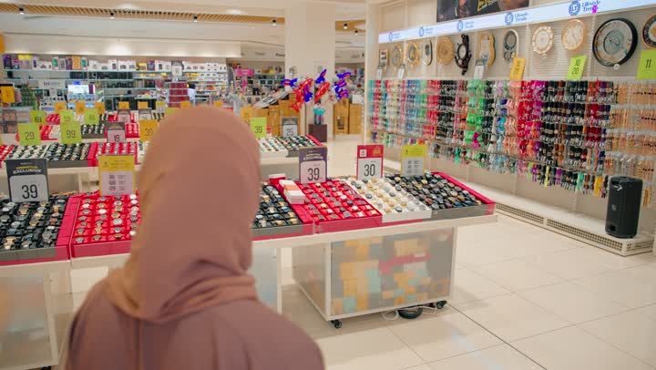 تصوير من الخلف لامرأة عربية خليجية اماراتية محجبة تسير داخل قسم عرض الساعات والإكسسوارات، مفهوم التسوق اليومي،دقة اختيار المنتجات، واهتمام المستهلك بمقارنة الخيارات قبل الشراء