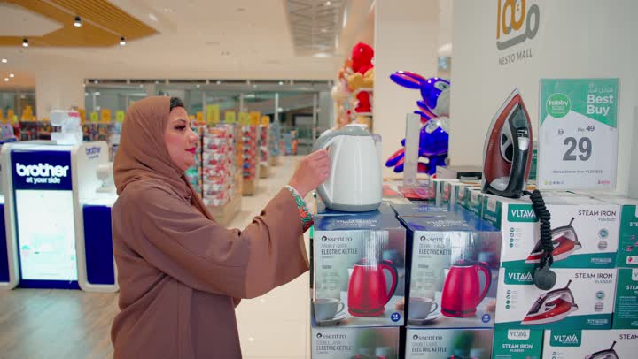 The consumer's interest in carefully choosing the right household tools, an Emirati Gulf Arab woman inside a store inspecting an electric kettle, ensuring the quality of the product before purchase, the concept of daily shopping.