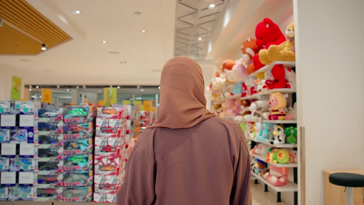 التأني في اختيار الألعاب الآمنة، مفهوم التسوق للأطفال، امرأة عربية خليجية اماراتية ترتدي الحجاب و العباءة داخل متجر مخصص لمنتجات الأطفال تقوم بتفقد لعبة دمية وتعاين شكلها وجودتها قبل الشراء