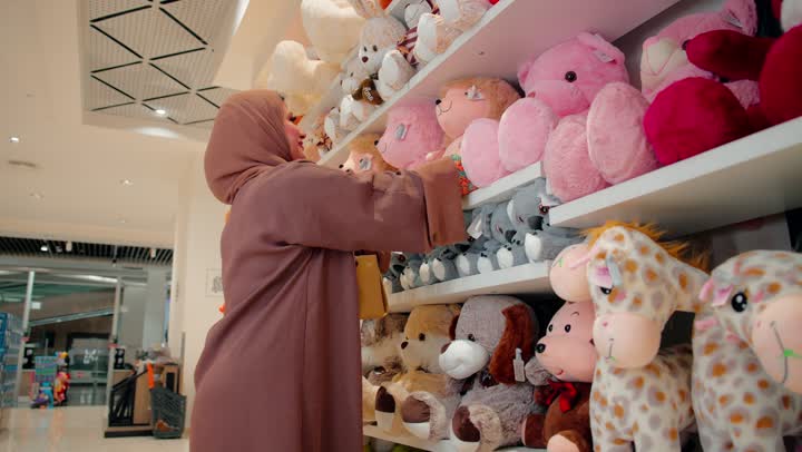 الاهتمام بانتقاء الألعاب الآمنة والمناسبة، مفهوم التسوق للأطفال، امرأة عربية خليجية اماراتية ترتدي الحجاب و العباءة داخل متجر مخصص لمنتجات الأطفال تقوم بتفقد لعبة دمية وتعاين شكلها وجودتها قبل الشراء
