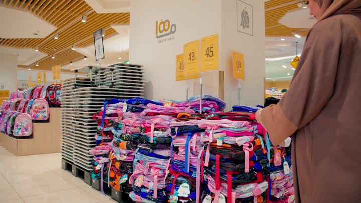 تقييم التصميم والجودة، اختيار احتياجات الأطفال المناسبة، الاستعداد للعودة إلى المدارس، امرأة عربية خليجية اماراتية محجبة ترتدي العباءة داخل متجر مستلزمات مدرسية تقوم بتفقد حقيبة مدرسية مخصصة للأطفال