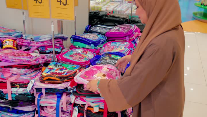 Choosing the appropriate needs for children, preparing for the return to school, inspecting the design and quality, an Arab Gulf Emirati woman wearing a hijab and an abaya inside a school supplies store is checking a school bag designed for children.