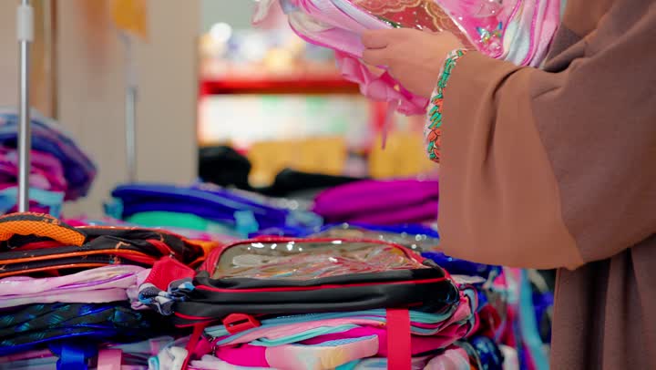 Carefully selecting children's supplies, preparing for back to school, inspecting design and quality, an Arab Gulf Emirati woman wearing a hijab and an abaya inside a school supplies store is checking a school bag designed for children.