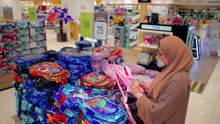 اختيار المستلزمات المناسبة للاطفال، الاستعداد للعودة إلى المدارس، معاينة التصميم و الجودة، امرأة عربية خليجية اماراتية محجبة ترتدي العباءة داخل متجر مستلزمات مدرسية تقوم بتفقد حقيبة مدرسية مخصصة للأطفال