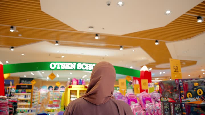 اختيار المستلزمات التعليمية المناسبة للأطفال، الاستعداد للموسم الدراسي، تصوير من الخلف لامرأة عربية اماراتية محجبة ترتدي العباءة داخل متجر مستلزمات مدرسية تسير بين الممرات وتتأمل الأقسام المختلفة المخصصة لمنتجات الأطفال والعودة إلى المدارس،عرض حقائب وأدوات مدرسية متنوعة