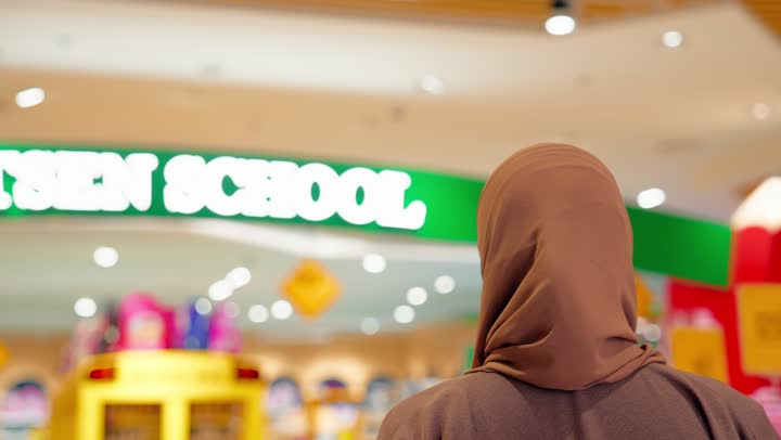 الاستعداد للموسم الدراسي، تصوير من الخلف لامرأة عربية اماراتية محجبة ترتدي العباءة داخل متجر مستلزمات مدرسية تسير بين الممرات وتتأمل الأقسام المختلفة المخصصة لمنتجات الأطفال والعودة إلى المدارس،عرض حقائب وأدوات مدرسية متنوعة، اختيار المستلزمات التعليمية المناسبة للأطفال