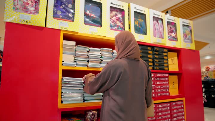 The concept of shopping for education, the importance of choosing the right books to develop children's skills, a rear view of an Emirati Gulf Arab woman wearing an abaya and hijab inside a school supplies store inspecting an educational book designed for children to learn English, reading the cover and content of the book before purchase, supporting the educational process.