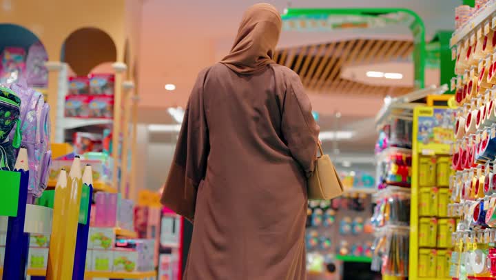 تنظيم الدراسة والعمل، اختيار الأدوات المكتبية، تصوير من الخلف لامرأة عربية خليجية اماراتية محجبة ترتدي العباءة و الحجاب داخل متجر تقف بجانب رف المستلزمات المكتبية والأدوات المدرسية، مفهوم التسوق اليومي