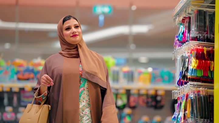 اختيار الأدوات المكتبية، امرأة عربية خليجية اماراتية محجبة ترتدي العباءة و الحجاب داخل متجر تقف بجانب رف المستلزمات المكتبية والأدوات المدرسية، مفهوم التسوق اليومي، تنظيم الدراسة والعمل