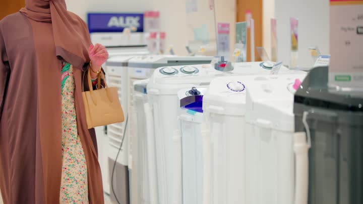 مفهوم التسوق الذكي، تصوير مقرب لامرأة عربية خليجية امارتية داخل متجر أجهزة منزلية تقوم بتفقد غسالة ملابس ومراجعة مواصفاتها قبل الشراء، اختيار الأجهزة المنزلية المناسبة
