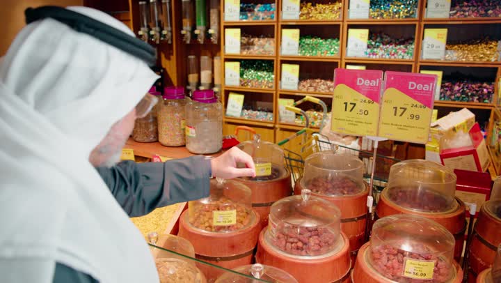 Arab food culture, the concept of conscious shopping, fresh food products, an Emirati Gulf Arab man wearing traditional attire inside a grocery store selecting dates and checking their quality, a healthy lifestyle, buying food products.