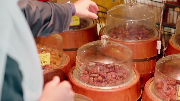 The concept of conscious shopping, fresh food products, an Arab Gulf Emirati man wearing traditional attire inside a grocery store selecting dates and ensuring their quality, Arab food culture, a healthy lifestyle, purchasing groceries.