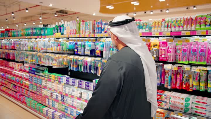 الاهتمام بالمظهر والنظافة الشخصية، مفهوم التسوق اليومي، رجل عربي خليجي اماراتي يرتدي الزي التقليدي يقف داخل سوبرماركت يقوم بقراءة عبوه منتج عناية شخصية من على الرفوف، متاجر البيع بالتجزئة