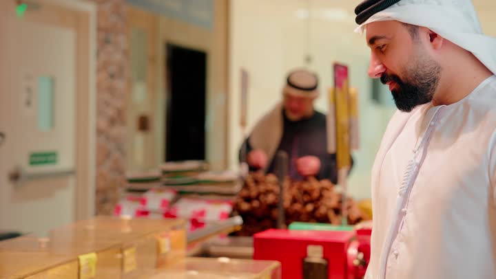رجل عربي خليجي اماراتي يرتدي الكندورة و الغترة في المتجر يقوم بشراء المكسرات والتسالي ، مفهوم الوجبة الخفيفة ، السلع والمواد الغذائية المختلفة ، مستلزمات المستهلك