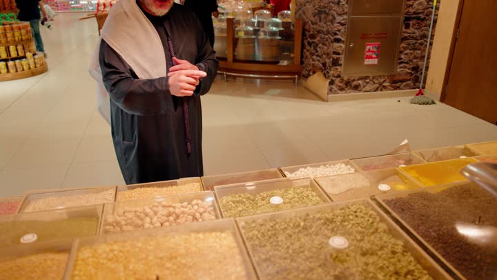 اختيار المنتجات الغذائية وتحديد الكمية المناسبة قبل الشراء،رجل عربي خليجي اماراتي مسن يرتدي الزي التقليدي يقف داخل متجر مواد غذائية تقليدي،  الاشارة إلى أصناف الحبوب والمكسرات المعروضة داخل عبوات شفافة