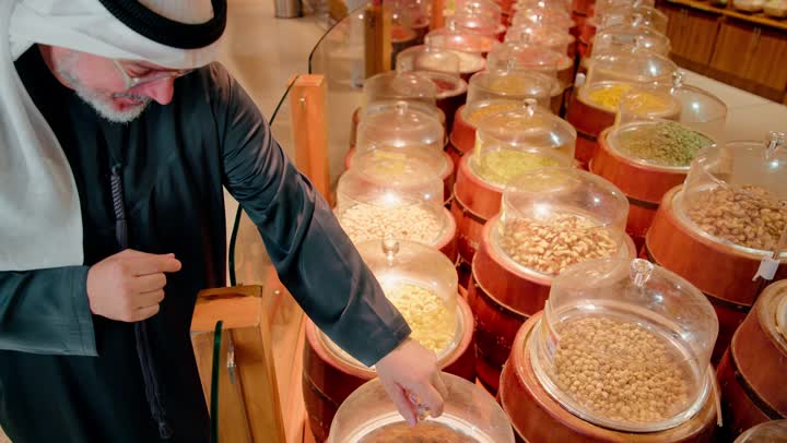 تفقد جودة المكسرات واختيار الكمية المناسبة، تصوير مقرب لرجل عربي خليجي اماراتي مسن داخل متجر مواد غذائية تقليدي يقوم باختيار مكسرات الكاجو من أوعية عرض شفافة، تسوق المنتجات غذائية