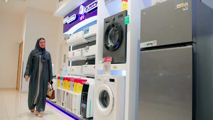 مقارنة التفاصيل الفنية قبل الشراء، اختيار الأجهزة المنزلية، امرأة عربية خليجية إماراتية محجبة تقف داخل متجر للأجهزة الكهربائية تتفقد ثلاجة منزلية وتقرأ بطاقة المواصفات المثبّتة عليها