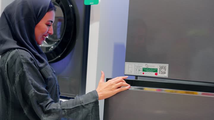 Choosing home appliances, a close-up shot of a veiled Emirati Gulf Arab woman standing inside an electrical appliances store inspecting a household refrigerator and reading the specifications label attached to it, comparing the technical details before making a purchase.