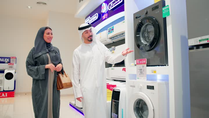 زوجان عربيان خليجيان اماراتيان يرتديان الزي التقليدي يقفان داخل متجر للأجهزة الكهربائية يقومان مناقشة ومقارنة التفاصيل قبل الشراء، مفهوم التسوق العائلي، تعاون واتخاذ قرار مشترك، اختيار ثلاجة مناسبة