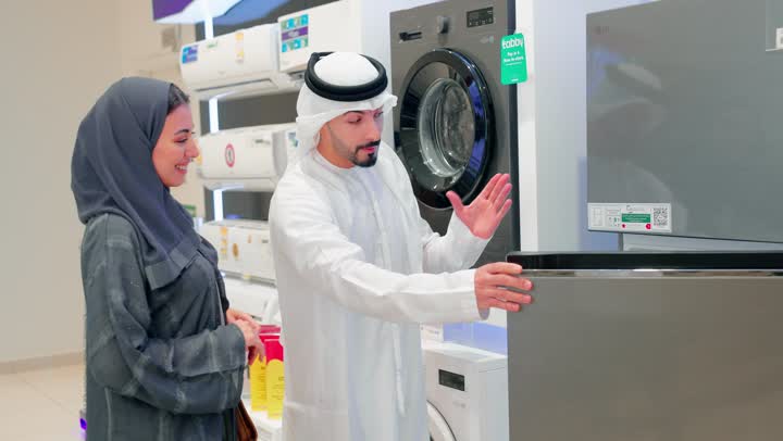 مفهوم التسوق العائلي، تعاون واتخاذ قرار مشترك، زوجان عربيان خليجيان اماراتيان يرتديان الزي التقليدي يقفان داخل متجر للأجهزة الكهربائية يقومان مناقشة ومقارنة التفاصيل قبل الشراء، اختيار ثلاجة مناسبة