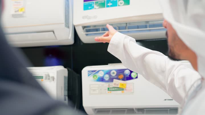 اختيار جهاز التبريد المنزلي، تصوير مقرب لرجل عربي خليجي يرتدي الزي التقليدي داخل متجر للأجهزة الكهربائية يشير إلى مكيف هواء جداري معروض على الحائط برفقة زوجته، مناقشة ومقارنة المواصفات الفنية قبل الشراء