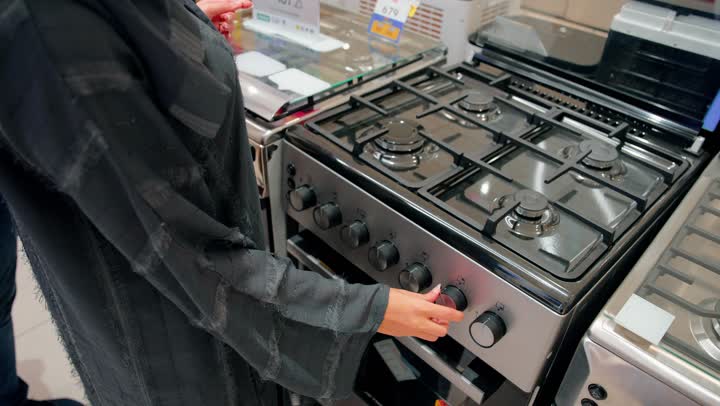 مفهوم التسوق للأجهزة المنزلية، فحص جودة الجهاز وملحقاته الداخلية، اختيار وشراء فرن كهربائي معروض، اقتناء أدوات المطبخ الحديثة، تصوير مقرب لامرأة عربية خليجية امارتية ترتدي العباءة تقف داخل متجر أجهزة منزلية، مقارنة المواصفات التقنية، تجهيز المنزل بأجهزة حديثة