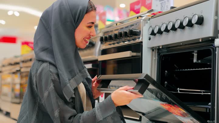 اختيار وشراء فرن كهربائي معروض، اقتناء أدوات المطبخ الحديثة، تصوير مقرب لامرأة عربية خليجية امارتية ترتدي العباءة تقف داخل متجر أجهزة منزلية، فحص جودة الجهاز وملحقاته الداخلية، مفهوم التسوق للأجهزة المنزلية، مقارنة المواصفات التقنية، تجهيز المنزل بأجهزة حديثة