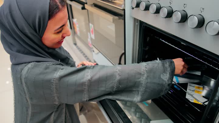 اقتناء أدوات المطبخ الحديثة، تصوير مقرب لامرأة عربية خليجية امارتية ترتدي العباءة تقف داخل متجر أجهزة منزلية ، اختيار وشراء فرن كهربائي معروض، فحص جودة الجهاز وملحقاته الداخلية، مفهوم التسوق للأجهزة المنزلية، مقارنة المواصفات التقنية، تجهيز المنزل بأجهزة حديثة