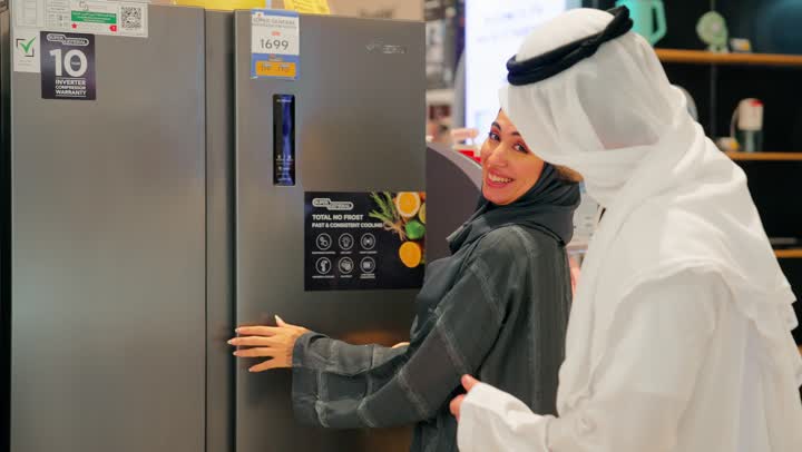 The concept of family shopping, discussing and comparing specifications before purchase, a close-up shot of an Arab Gulf couple wearing traditional attire standing in front of display refrigerators inside an electrical appliances store, choosing a suitable home refrigerator, collaboration and making a joint decision.