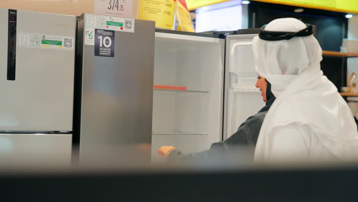 مناقشة ومقارنة المواصفات قبل الشراء، تصوير مقرب لزوجان عربيان خليجيان يرتديان الزي التقليدي يقفان أمام ثلاجات عرض داخل متجر للأجهزة الكهربائية، اختيار ثلاجة منزلية مناسبة، مفهوم التسوق العائلي، تعاون واتخاذ قرار مشترك
